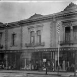 Image: a black and white photograph of a two storey terrace building with large plate glass shop windows on the first floor displaying a range of clothing and fabrics