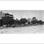 Image: dozens of men in early 20th century working clothes stand along a dirt road lined with single and two storey buildings and mature trees laying a telegraph cable.