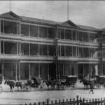 Image: A line of horse drawn vehicles waits outside a huge three storey hotel with balconies on the second and third floors and a verandah on the ground floor.