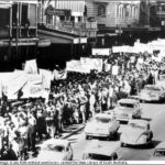 Image: a large group of people holding protest signs against the Vietnam War march down a city street