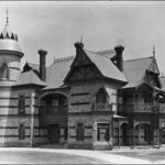 Image: A large, two-storey stone mansion with a cylindrical turret comprising one corner of the structure