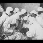 Image: A group of surgeons and medical technicians in white surgery gowns surround a patient on an operating table. Three men closest to the patient are performing an operation