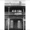 Image: a group of five women in white dresses in an early 20th century style stand on a balcony of a two storey building which has signs declaring it the Globe Dining Rooms