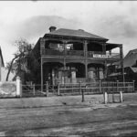 Image: a two storey building with a combination verandah and balcony, decorated with lattice work, sits behind a wooden fence. Washing dries on a line on the balcony.