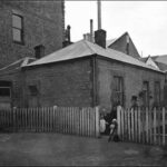 Image: A family in Victorian attire stand in front of a rectangular, single-storey stone and brick building. A white picket fence surrounds the building, and a few multi-storey stone and brick structures are visible behind it