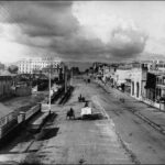 Image: dark clouds hang over a wide dirt city street which is lined with a variety of buildings from single storey tin sheds to large stone constructions of up to six storeys. Travelling down the road are a range of horse drawn vehicles.