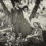 Image: A moustachioed man in a suit sits on a rug. Leaning against a tree behind him are a young man, young woman, and and adult woman in Edwardian attire