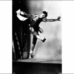 Image: A man in stage make-up and costume dances on a stage with arms outstretched