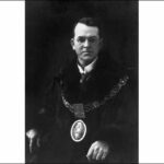 Image: A man with receeding dark hair and glasses poses for a portrait while wearing a dark robe and livery collar