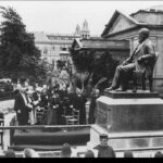 Image: A crowd of people in Edwardian-era attire gather around a large bronze statue of a bearded man sitting in a chair