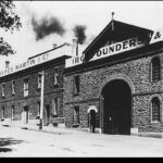 Image: A large complex of stone two-storey buildings next to a street. A sign on the front of the building reads ‘James Martin & Co., Iron Founders’