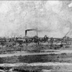 Image: Smoke billows from the chimney of a low building surrounded by mining equipment