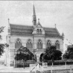 Image: a two storey gothic style building with arched church style windows and an arched portico protruding from the front, providing a balcony above. A thin spire rises from the centre of the cross gable roof.