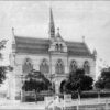 Image: a two storey gothic style building with arched church style windows and an arched portico protruding from the front, providing a balcony above. A thin spire rises from the centre of the cross gable roof.