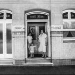 Image: a black and white shot of a smiling older lady holding hands with a toddler, and a tween, in the entrance to a private lounge