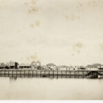 Image: A line of buildings sit atop a long wharf. In front of the wharf is a river, in which three small boats are moored