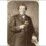 Image: An old photograph of a man wearing a dark coat, bow-tie and medals posing with one hand holding a book upright on a table by his side, the other, his right, tucked in between the buttons of his coat