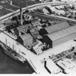 Image: An aerial photograph of a complex of large, multi-storey brick buildings and corrugated metal structures located next to a river. Two sailing ships are moored alongside a wharf associated with the complex