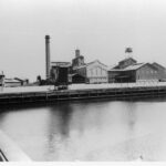 Image: A complex of multi-storey brick buildings and large, corrugated metal-clad warehouses fronted by a river. A large brick chimney is visible at one end of the complex