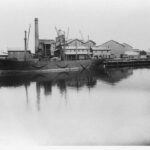 Image: A complex of multi-storey brick buildings and large, corrugated metal-clad warehouses fronted by a river. A large brick chimney is visible at one end of the complex, and a hulk is moored against its wharf