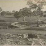 Image: A large paddock featuring a dirt road that crosses a creek via a stone bridge. A large house is just visible in the background