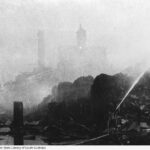 Image: A brick chimney and a few brick multi-storey buildings are shrouded in smoke from a fire. The burning sugar stock is visible in the foreground