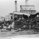 Image: Destroyed and partially-destroyed buildings are visible behind a large, burnt pile gunny sacks interspersed with twisted sheets of corrugated metal