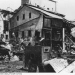 Image: A burned-out corrugated metal building stands next to the partially-destroyed wall of a multi-storey brick building. Sheets of twisted corrugated metal and other debris are visible in the foreground