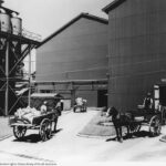 Image: Horse-drawn carts containing bags of sugar travel into a large corrugated metal-clad warehouse, while empty carts are transported out of another