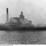 Image: A brick chimney and a few brick multi-storey buildings are shrouded in smoke from a fire. Other burned out structures are visible nearby