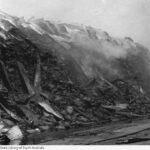 Image: A large, smouldering pile of charred gunny sacks, atop which are fragments of burned corrugated metal sheeting