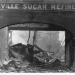 Image: A brick archway with the words ‘Glanville Sugar Refinery’ painted at its top is visible in front of a pile of burned, collapsed corrugated metal sheeting