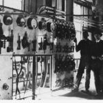 Image: Two men in 1920s attire stand inside a cavernous warehouse next to a large electrical switchboard