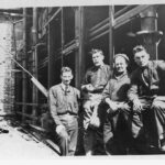 Image: A group of four men pose for a photograph outside a multi-storey brick building under construction