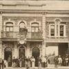 Image: A group of at least 24 men in 1880s attire stand on the street outside a two storey stone terrace building. A further two figures can be seen on the balcony above. The building features decorative window surrounds and extends over a large archway.