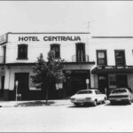 Image: a two storey corner hotel, painted white with dark windows and doors and a flat roof. Small, round balconetes protrude from some of the upper floor openings. 1980s era cars are parked on the street outside.