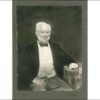 Image: a photograph of a seated, white haired man with a beard. His left hand is resting on a table and his right is on his lap. He is wearing grey trousers, a white shirt and waistcoat and a black jacket and bow tie.