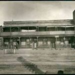 Image: a series of four terraced cottages amalgamated into a single boarding house. The building features a verandah/balcony combination and a low, solid parapet. Men and women stand in the doorways of each cottage behind metal fences and gates.