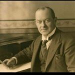 Image: A photographic portrait of a middle-aged, clean-shaven man in a suit sitting at a table