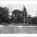 Image: A large park containing several trees is bordered by an iron fence. A dirt street and three lampposts are visible in the foreground