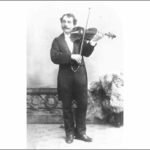 Image: A young moustachioed man in formal attire poses with a violin for a full-length photographic portrait