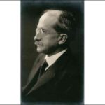 Image: A photographic portrait of a moustachioed man in a suit and wire-rimmed spectacles. He is shown in partial profile