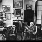 Image: An elderly bearded man sits in a chair and reads a book. The room contains a variety of furnishings and several paintings hang on the walls