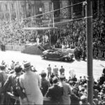 Image: A large crowd watches as a man escorts a woman down a set of stairs to a waiting car of 1950s vintage