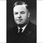 Image: A photographic head-and-shoulders portrait of a middle-aged Caucasian man with dark hair. He is clean-shaven and wearing a 1930s-era suit and tie