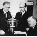 Image: Three middle-aged Caucasian men in mid-20th century suits examine a piece of scientific apparatus on a table