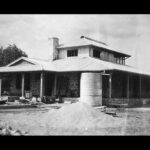 Image: A single-storey stone building with gabled roof and wraparound verandah. A corrugated metal water tank and evidence of construction is visible in the foreground