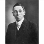 Image: A photographic head-and-shoulders portrait of a young man wearing a suit and wire-rimmed spectacles