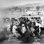 Image: A group of Afghan men and their camels stand in a remote area. A small tent is visible in the background