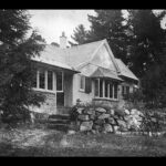 Image: A single-storey cottage nestled amongst several large cedar trees. The front of the house is bordered by a low stone wall